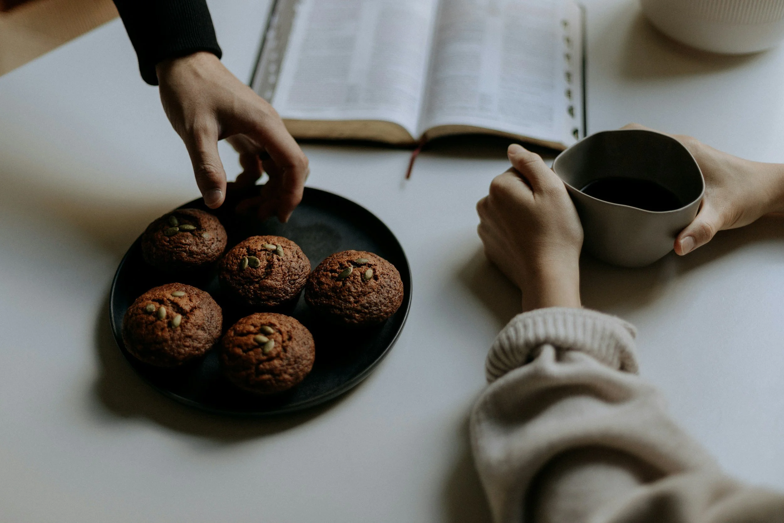 Plate of breakfast muffins with seeds, hands holding a cup, open book on table.