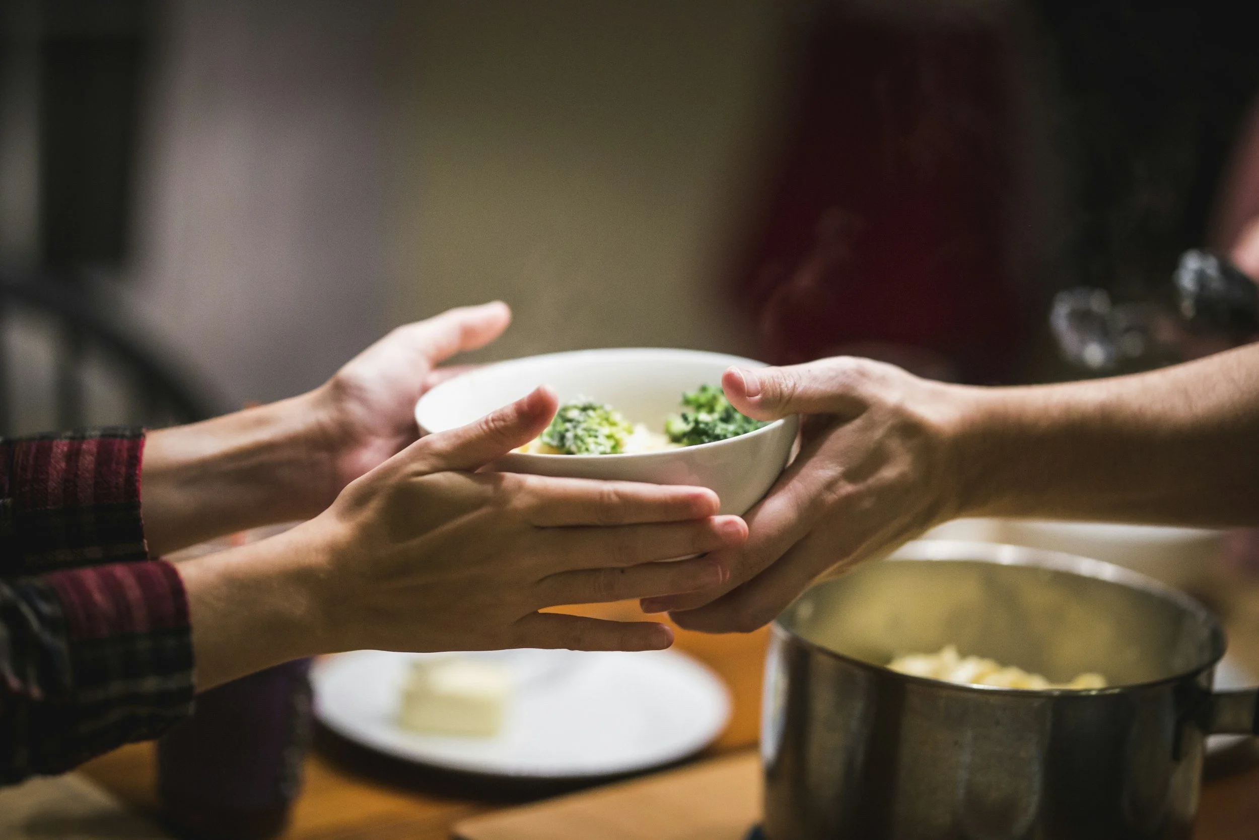 Two people exchanging a bowl of food over a table. A pot is visible on the table.
