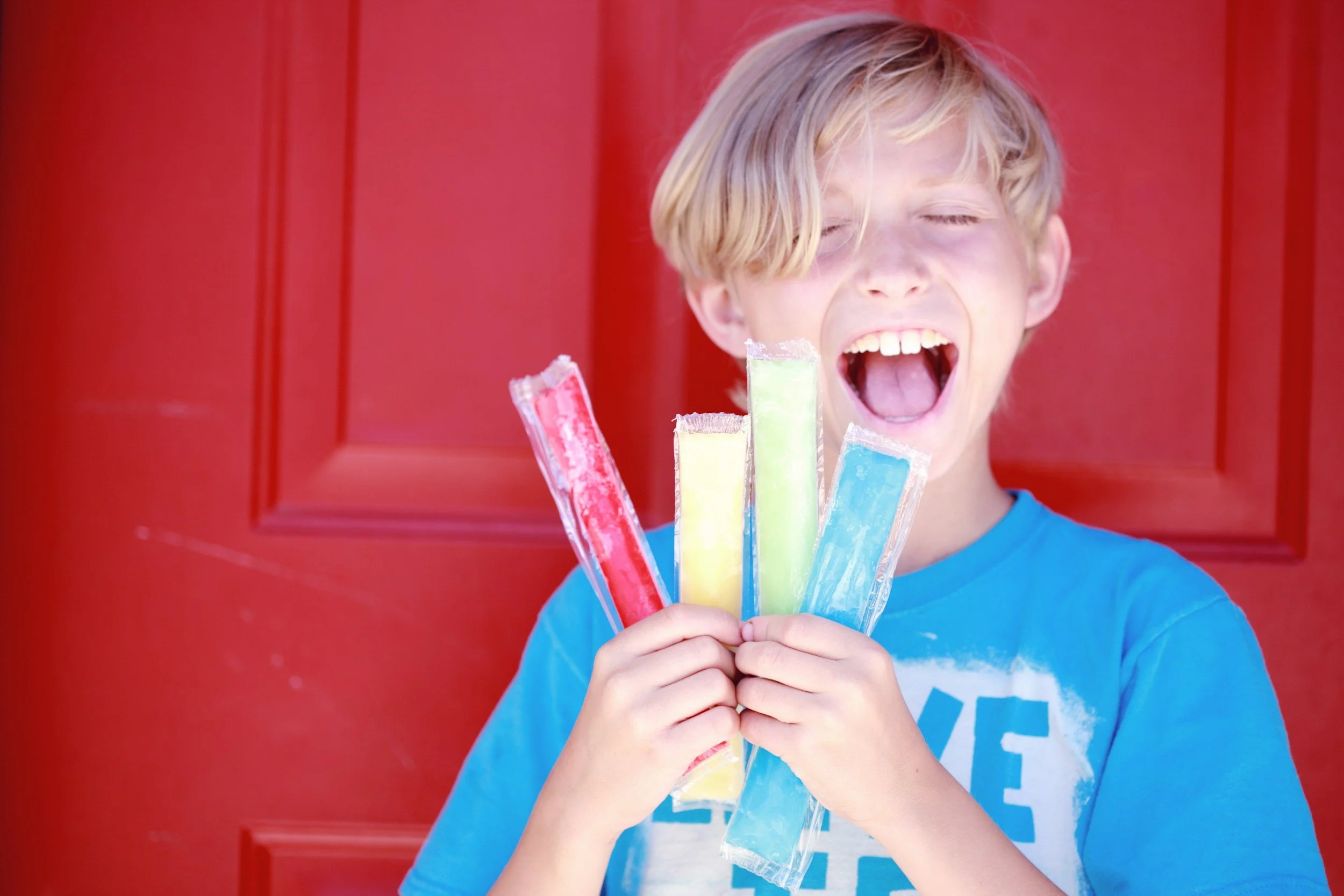 A boy with blonde hair, wearing a blue shirt, holding four colorful popsicles, smiling with eyes closed, standing in front of a red door.